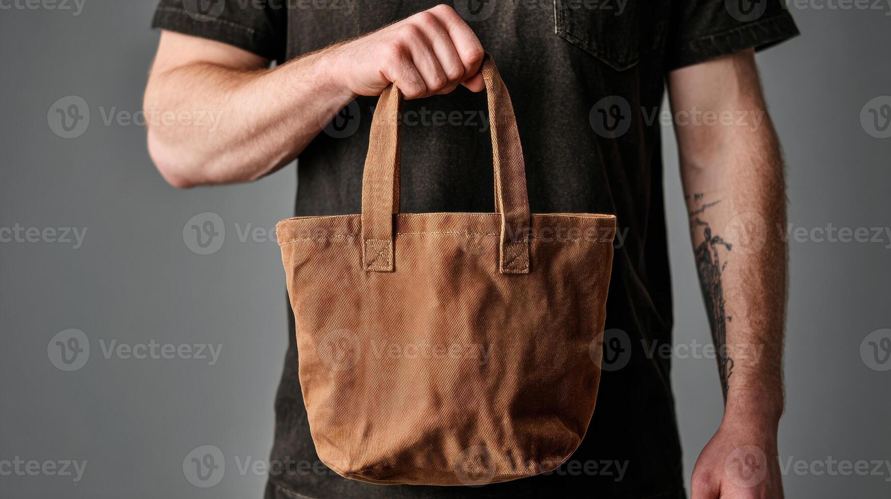 Hand Holding a Brown Canvas Tote Bag Against a Grey Background With Focus on the Bag and Part of a Mans Arm photo