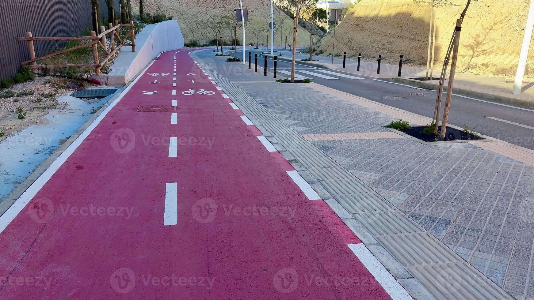 A pink bike lane winds through a bustling urban area, next to a landscaped path filled with lush trees photo