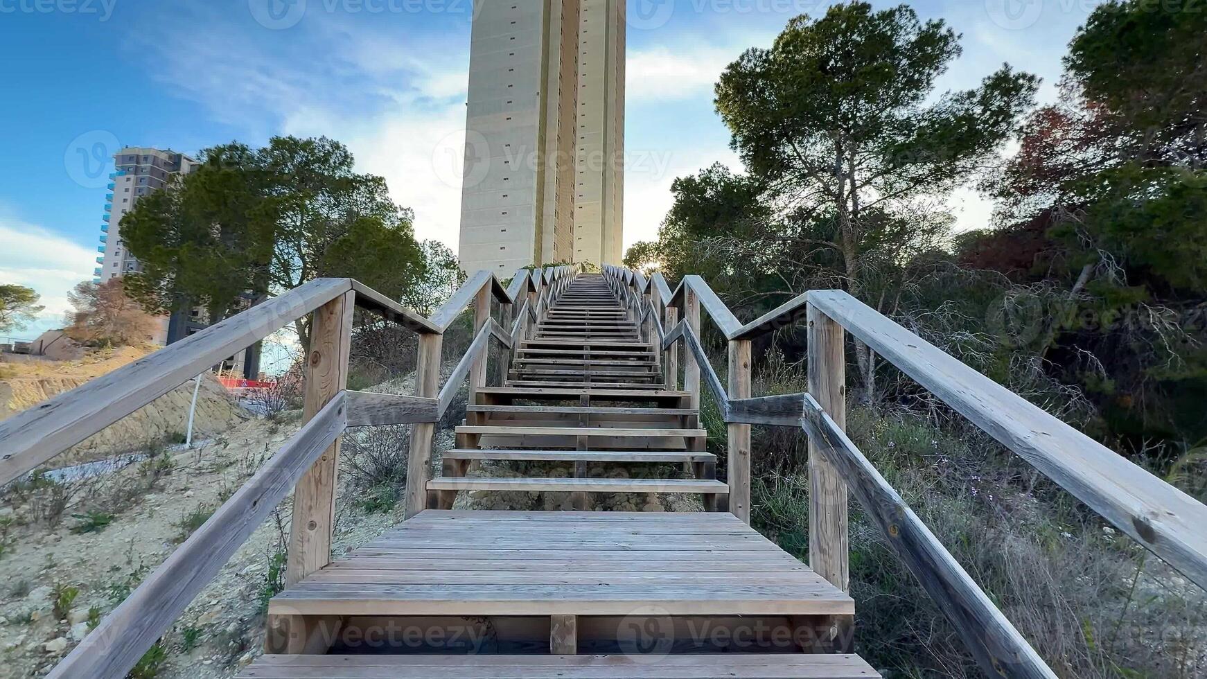 A beautifully crafted wooden staircase gracefully ascends towards a tall and impressive tower, surrounded by lush trees photo