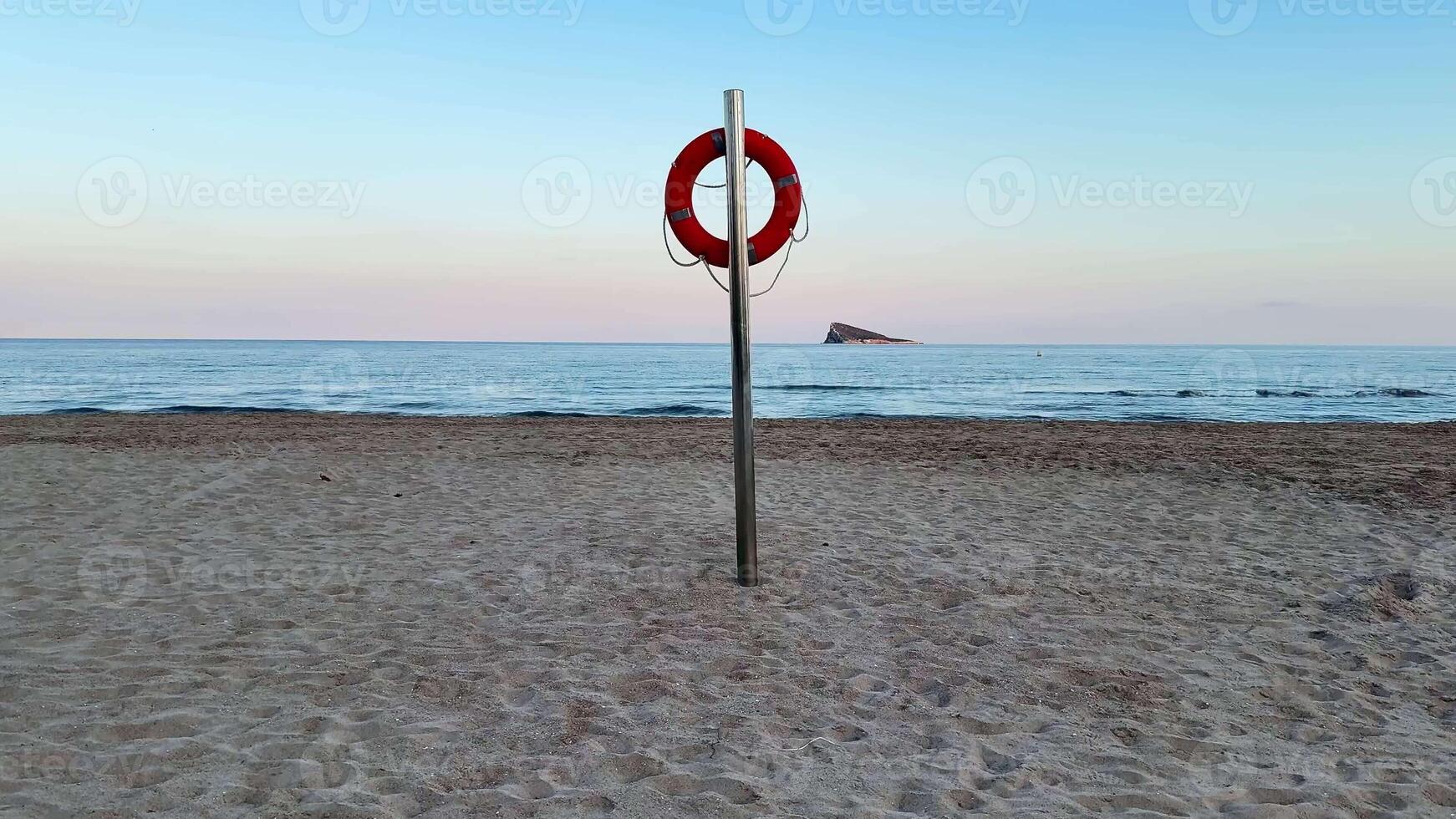 A bright lifebuoy stands tall on a serene beach as the sun slowly sets over the calm and peaceful sea photo
