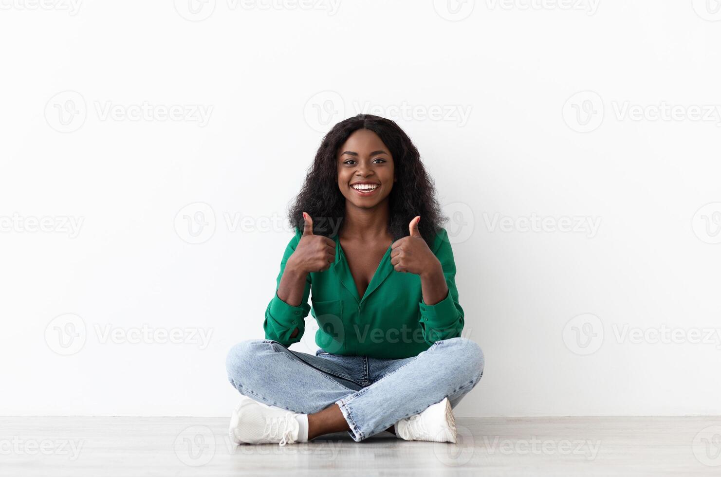 Happy african american young woman showing thumb ups, sitting on floor next to white wall, copy space, pretty black lady recommending something nice, enjoying her life, choice photo