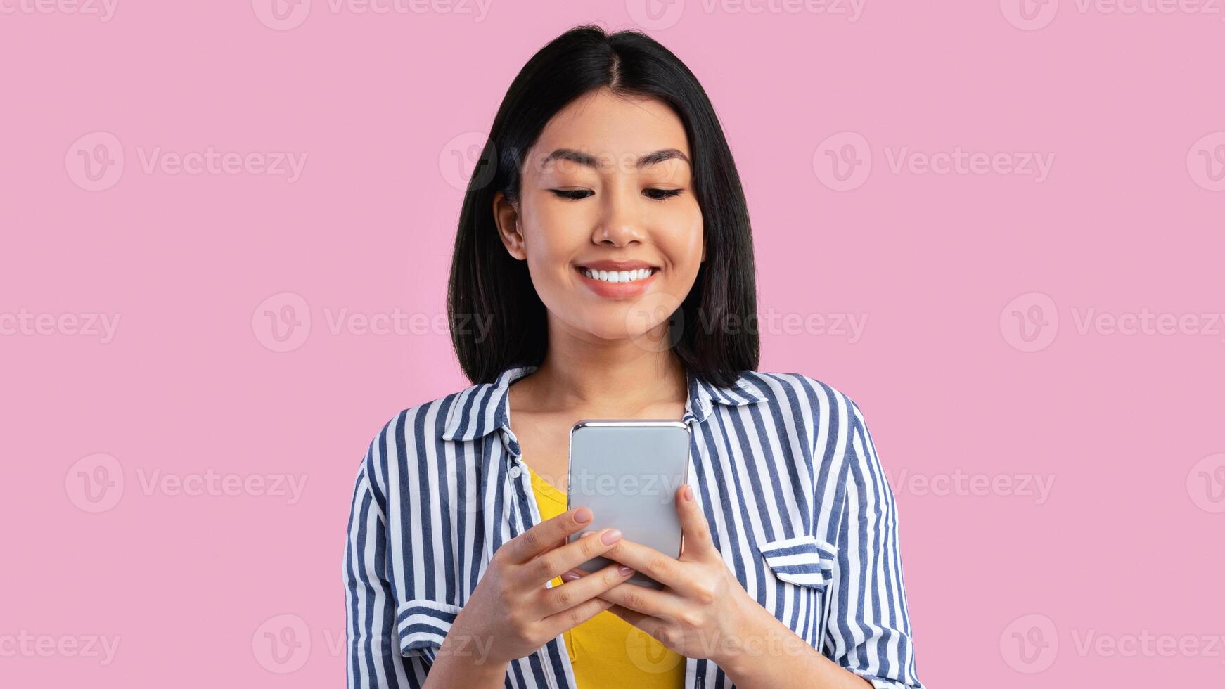 Amazing Application. Front portrait of smiling young asian woman using smartphone standing isolated over pink studio background. Cheerful lady texting sms message, chatting with friends, banner photo