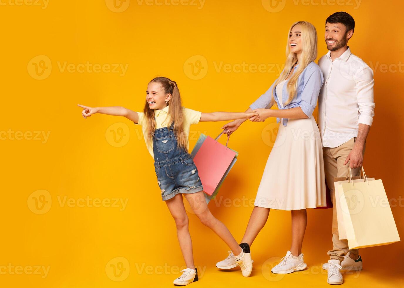 Family Shopping Concept. Daughter Pointing Finger Showing Something To Her Parents Over Yellow Studio Background. Free Space photo