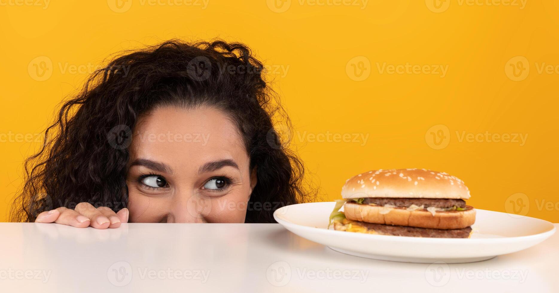 Funny Hungry Woman Looking At Burger On Plate, Peeking Out Table Posing Over Yellow Orange Studio Background. Dieting, Cheat Meal And Unhealthy Junk Food, Overeating Habit Concept. Panorama Banner photo