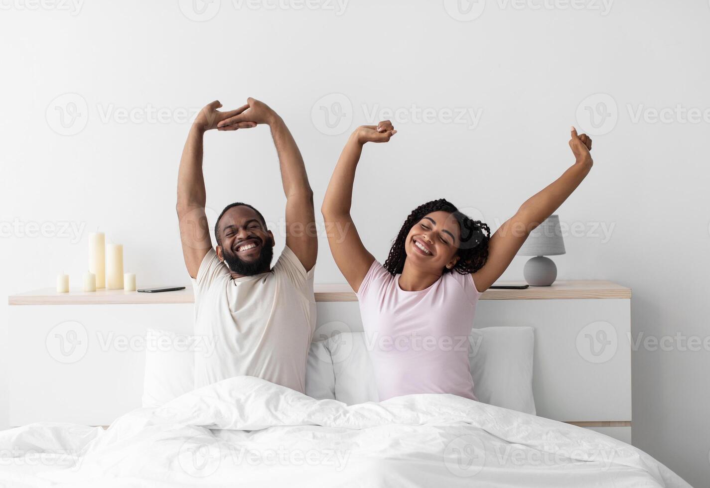 Happy millennial black husband and wife woke up, stretching bodies on white bed in minimalist bedroom interior. Couple in white t-shirts in morning after wake up. Healthy sleep, vacation and weekend photo