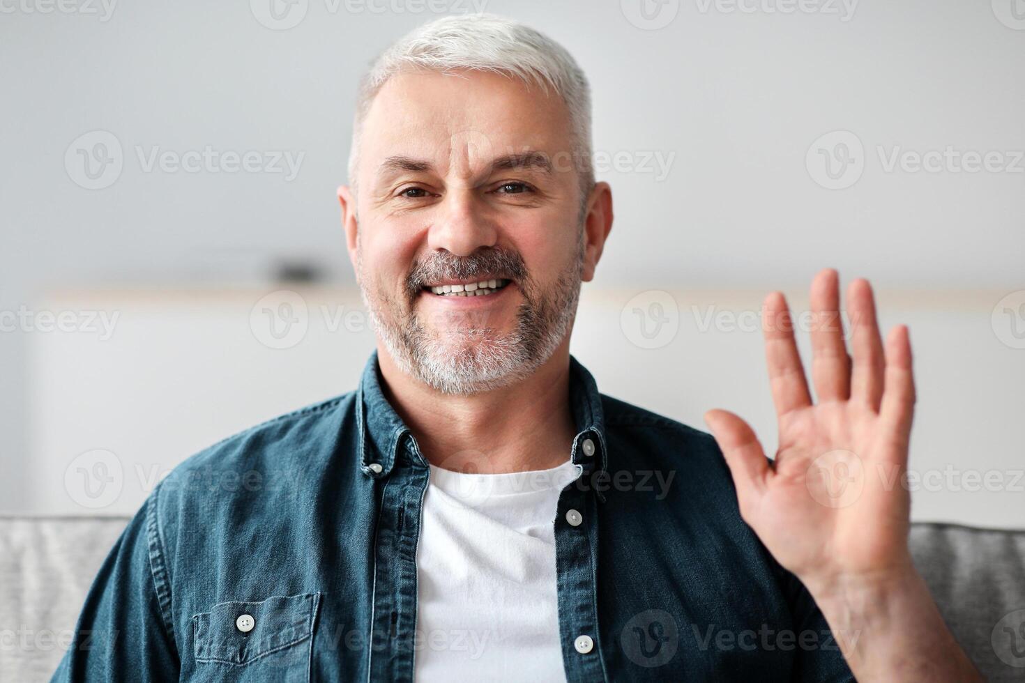 Handsome senior man having conference, smiling and waving at camera, device screen with telecommunication software frame. Elderly man having online party during COVID-19 pandemic photo