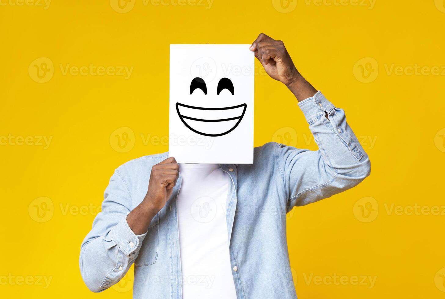 Happiness and success concept. Black male hiding face behind sheet of paper with drawn happy emoticon over yellow background in studio, copy space photo