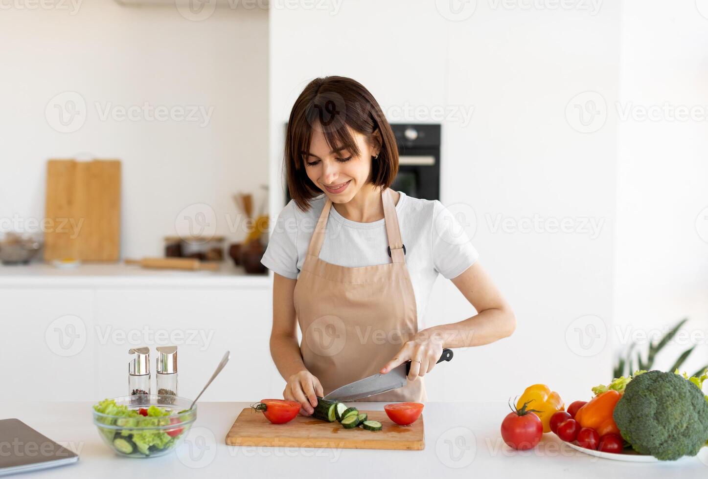 Young housewife cooking delicious and healthy food, preparing vegetable salad, slicing cucumber with knife on cutting board, standing in modern kitchen at home photo