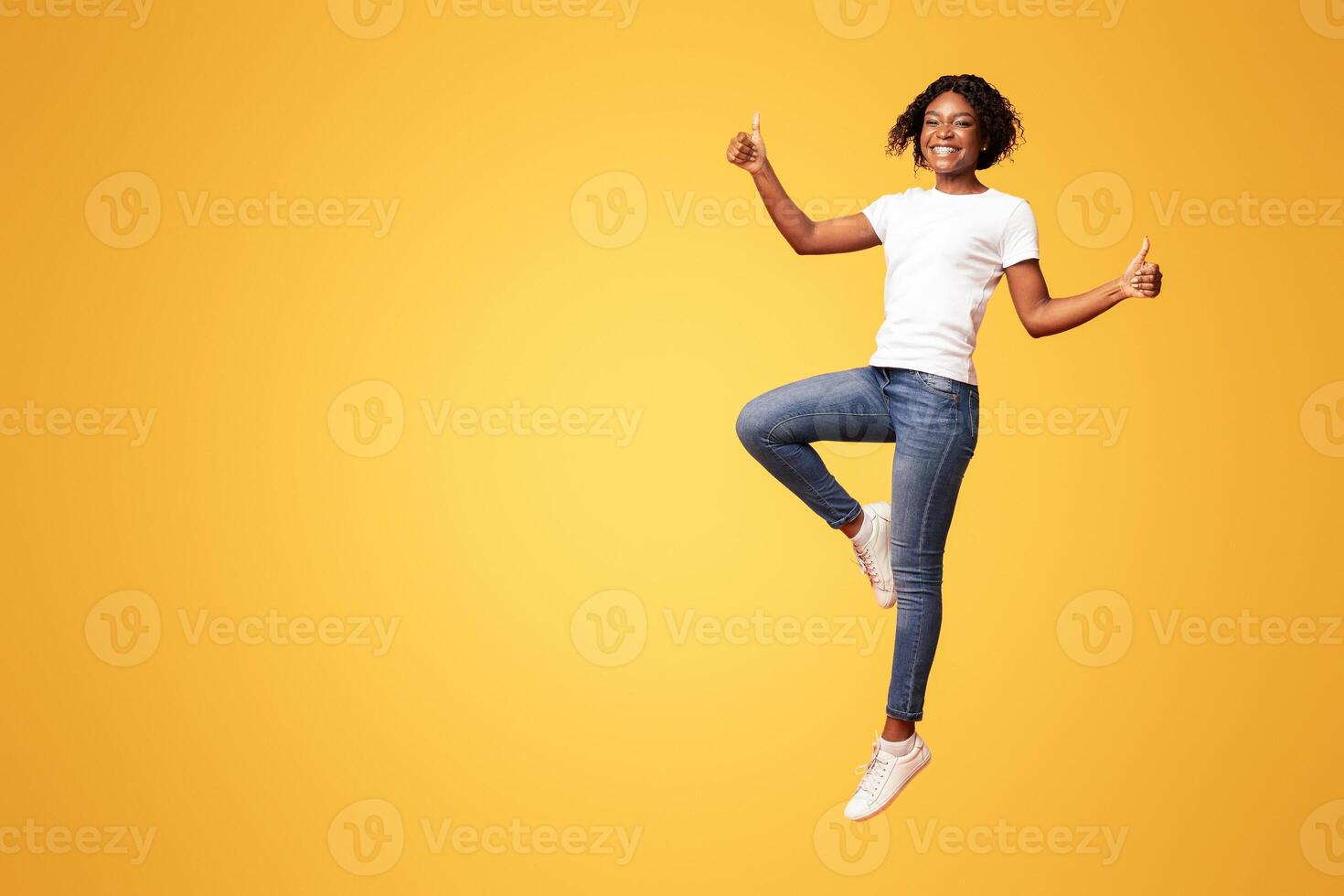 Carefree african-american millennial woman jumping up and showing thumb up over orange studio background, recommending something, celebrating success, showing her happiness, full length shot photo