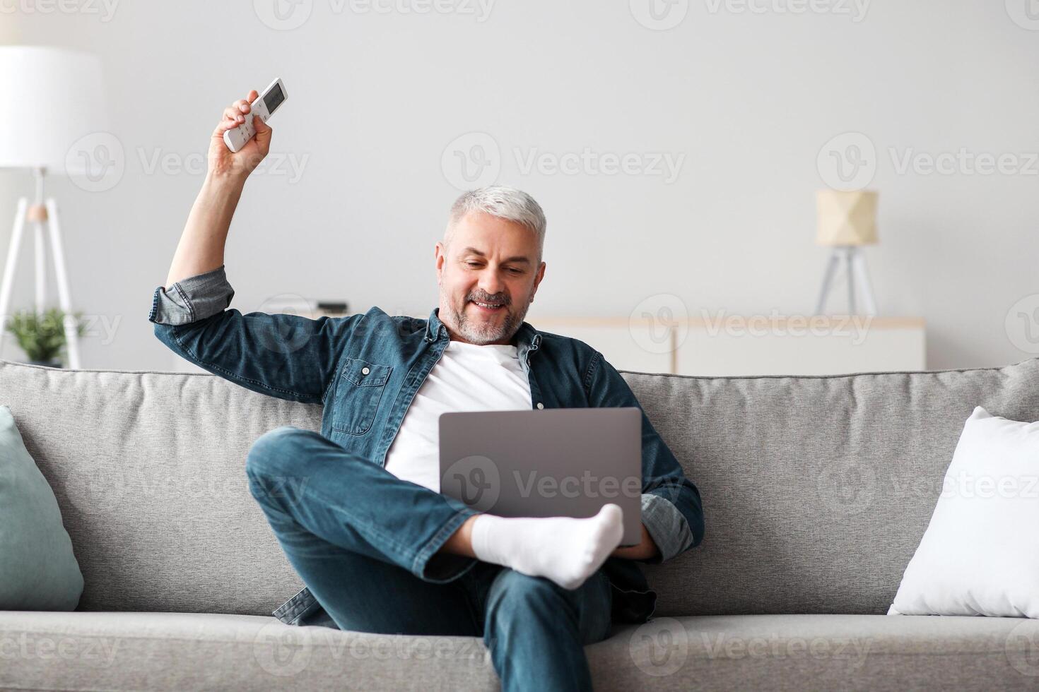 Relaxed mature man sitting on couch with laptop, turning on AC, feeling hot. Happy senior man enjoying time at home, surfing on Internet, using notebook, holding remote for air conditioner, copy space photo