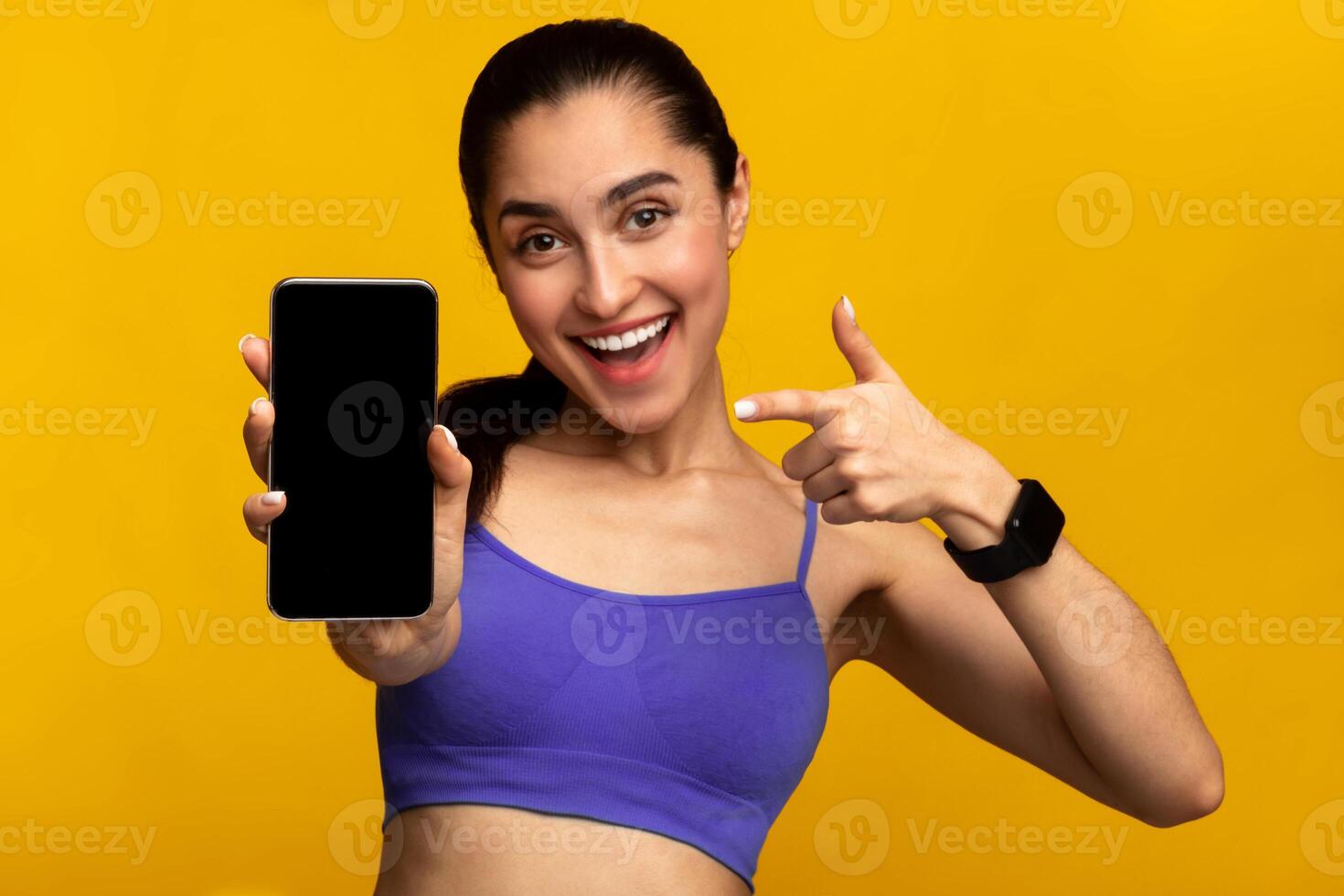 Sports App. Portrait of excited fit young lady holding and showing smartphone, pointing and indicating black empty screen for mockup template, wearing fitness tracker, orange studio background photo