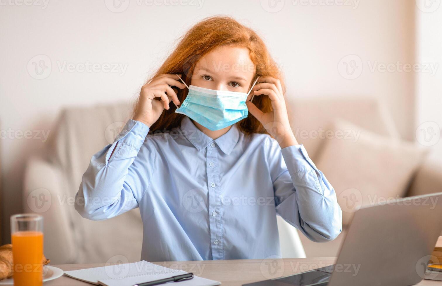 Remote Education, Quarantine, Lockdown, Protection. Portrait of girl wearing medical face mask studying at home on laptop computer, sitting at table and looking at camera, doing homework photo