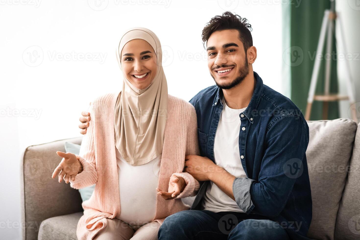 Portrait Of Joyful Muslim Couple Awaiting Baby Posing At Home, Happy Pregnant Islamic Woman In Hijab Relaxing On Couch With Her Loving Husband, Enjoying Future Parenthood, Copy Space photo
