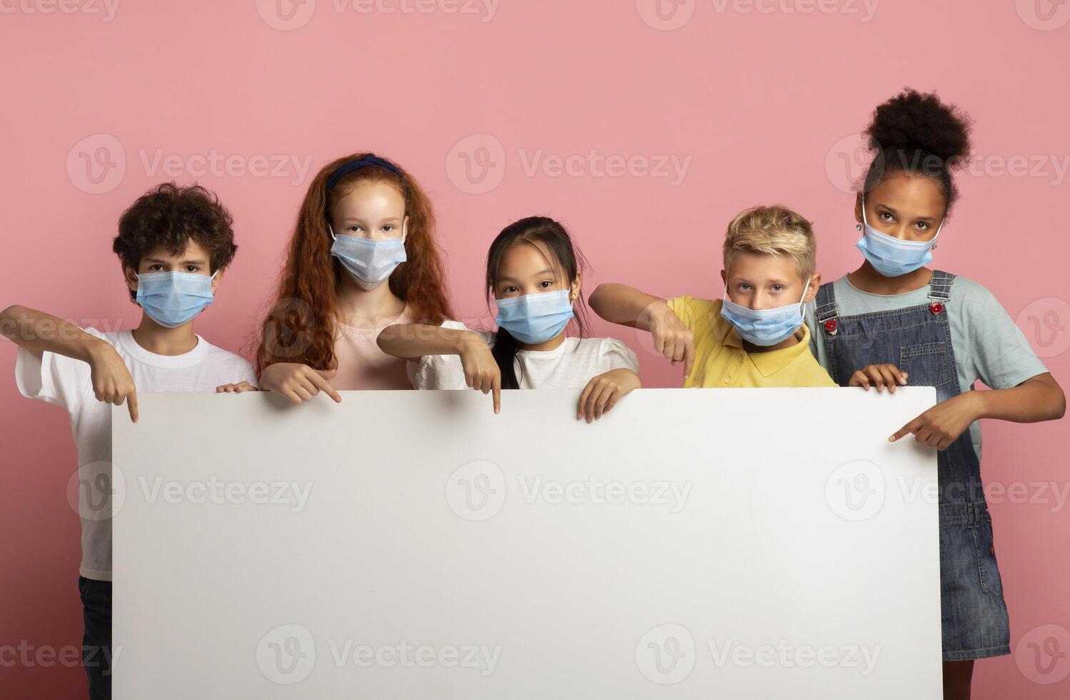 Multiethnic group of kids in medical masks pointing at blank banner over pink background, mockup for design photo