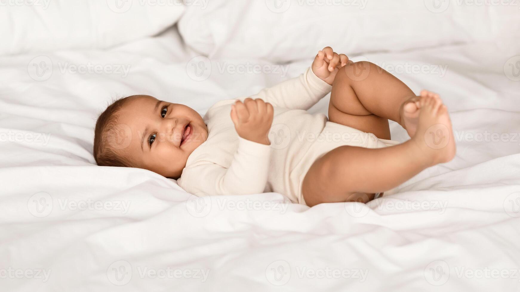 In The Morning. Portrait of adorable smiling black infant lying on the back in bed on white bedsheet at home. Positive healthy laughing afro child wearing bodysuit, looking at camera. Happy Childhood photo