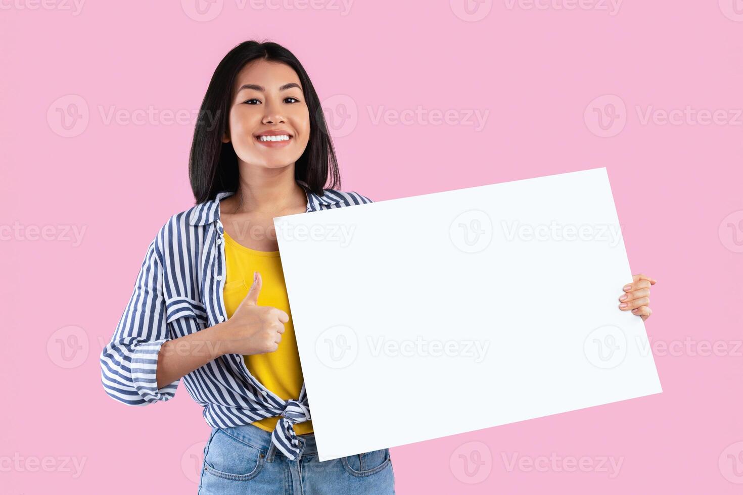 Portrait Of Cheerful Asian Woman Showing Thumbs Up Sign Gesture And Holding Blank Paper Poster For Your Text Standing With White Square Board For Advertising Isolated Over Pink Studio Background photo