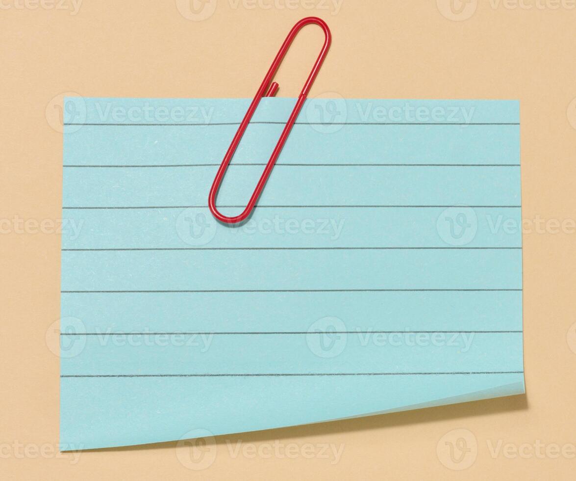 Blue sticky note with a red paper clip attached to it, placed on a beige background. Top view photo