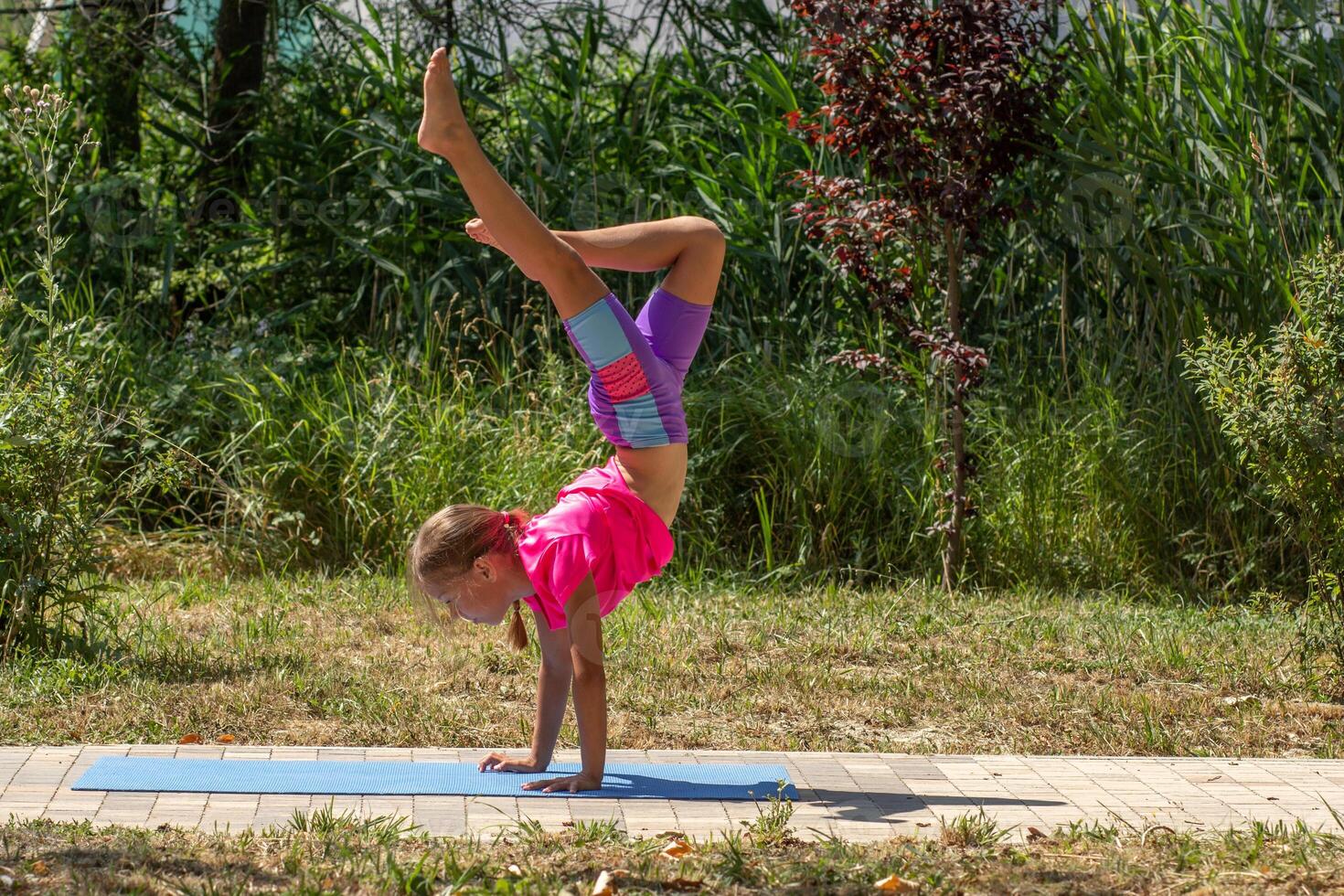 A young girl practices a dynamic handstand on a yoga mat outdoors, supporting her weight on both hands while one leg is extended upward and the other bent. photo