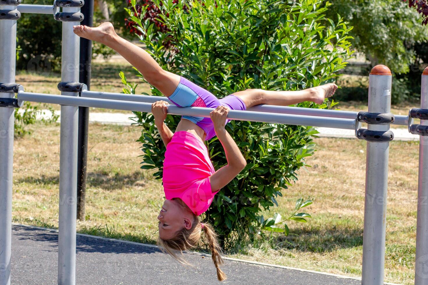 A child hangs upside down from a horizontal bar, legs extended in a full split, demonstrating impressive flexibility and body control while suspended in the air. photo