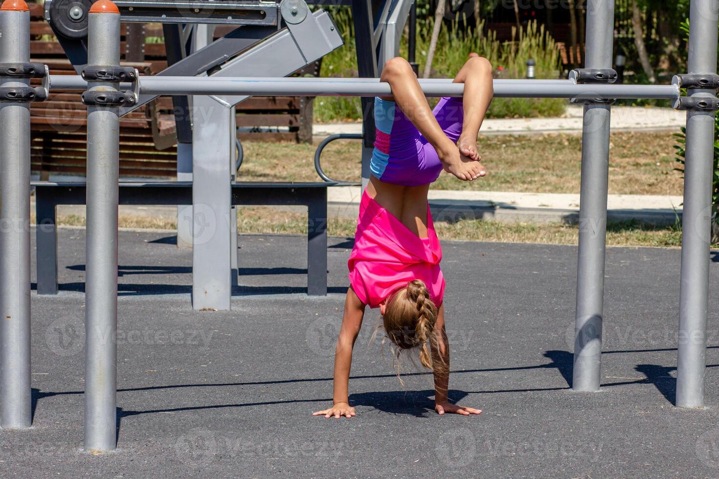 A child balances upside down on a horizontal bar, supporting the body with bent arms while the legs are lifted and bent overhead. The pose shows strength, coordination, and gymnastic control. photo