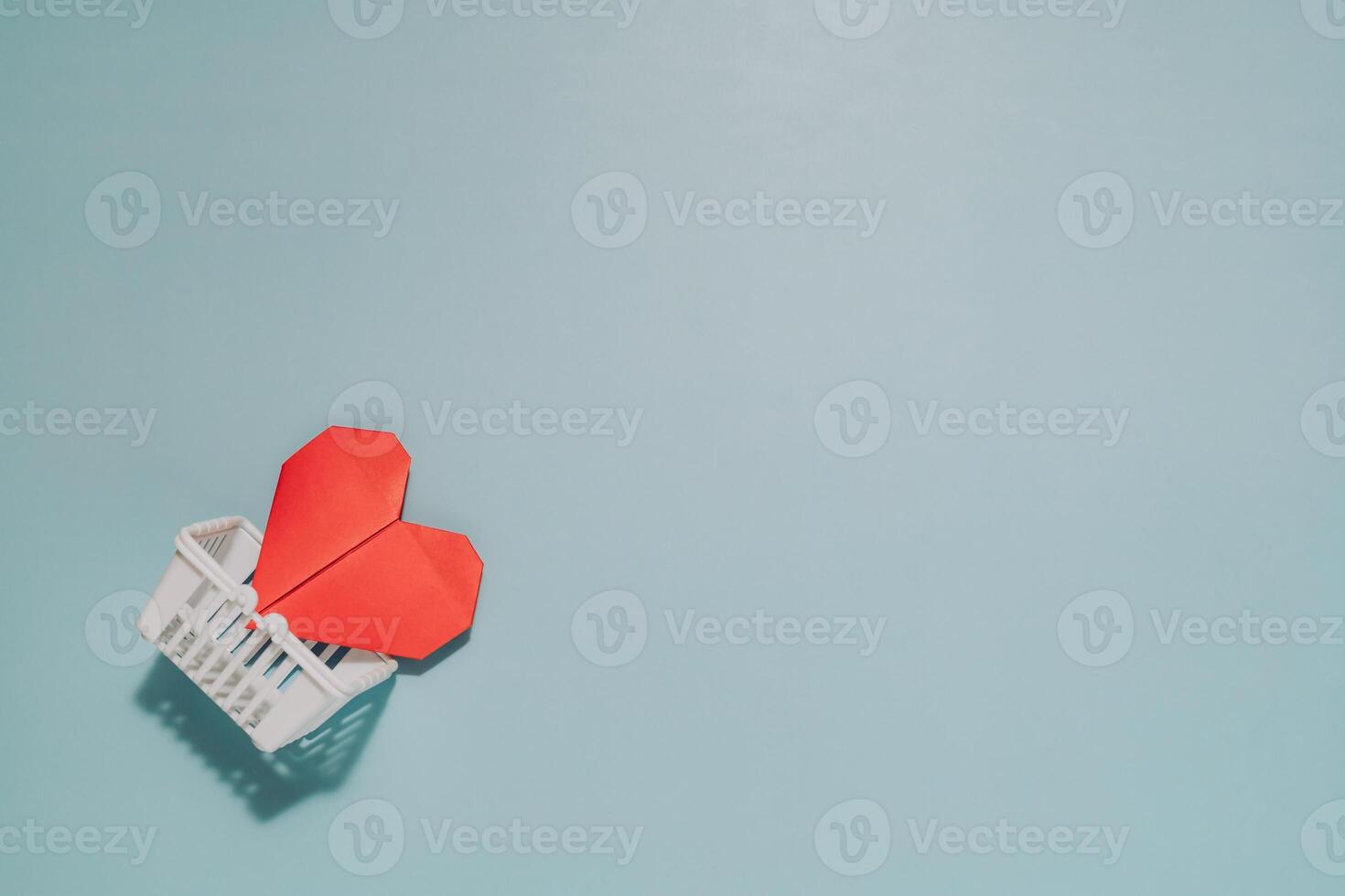 Bright red origami heart in a white miniature shopping basket on muted blue. Symbolizes self love, choosing health, giving love, or shopping for care. photo