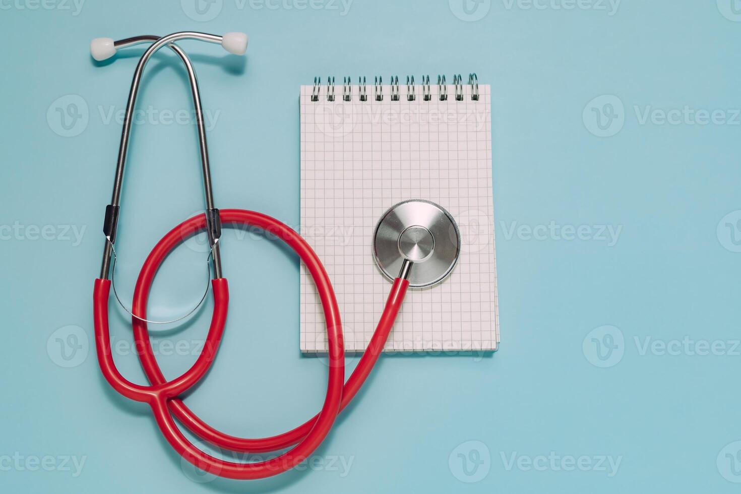 Vibrant red stethoscope next to an open spiral notepad with grid paper, all on light blue. Ideal for medical consultation, patient records, diagnosis, and healthcare planning. photo