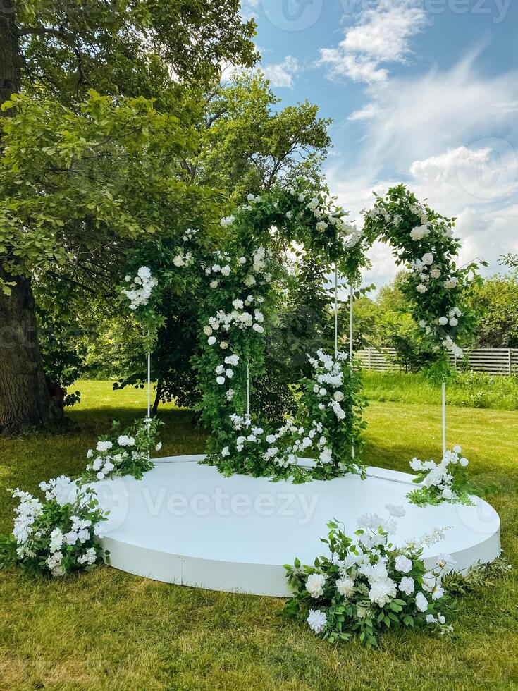 In a beautiful park, there is a stunning wedding arch that is elegantly decorated with gorgeous white flowers and lush greenery, creating a picturesque setting for a lovely celebration photo