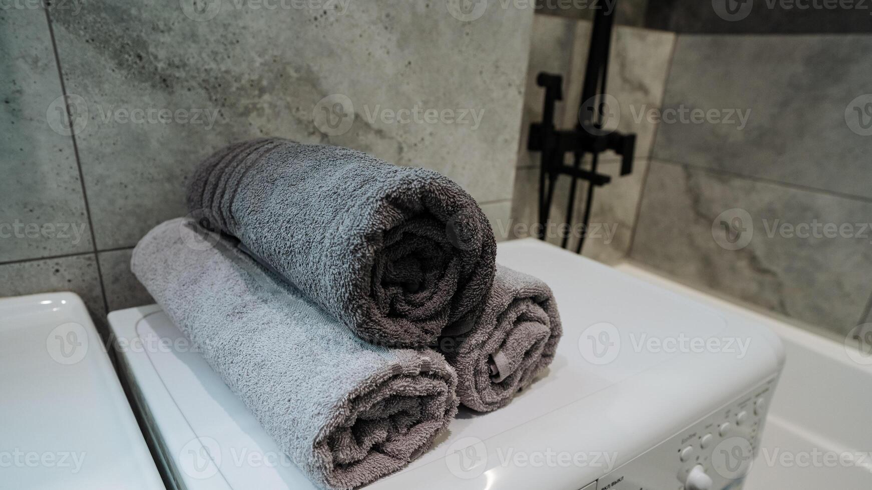 In a small bathroom, three towels are neatly rolled up and placed on top of a washing machine, surrounded by various household supplies and plumbing fixtures, adding to the decor photo