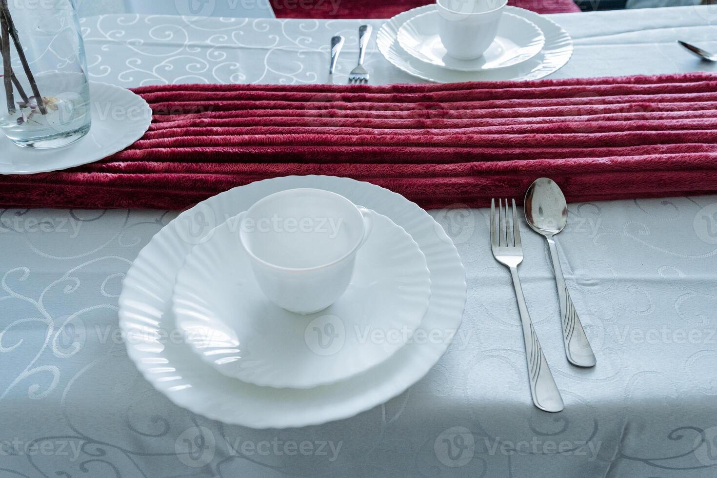 A beautifully arranged dining table showcasing elegant white plates and polished silverware, all complemented by a striking red table runner that adds a vibrant touch to the setting photo