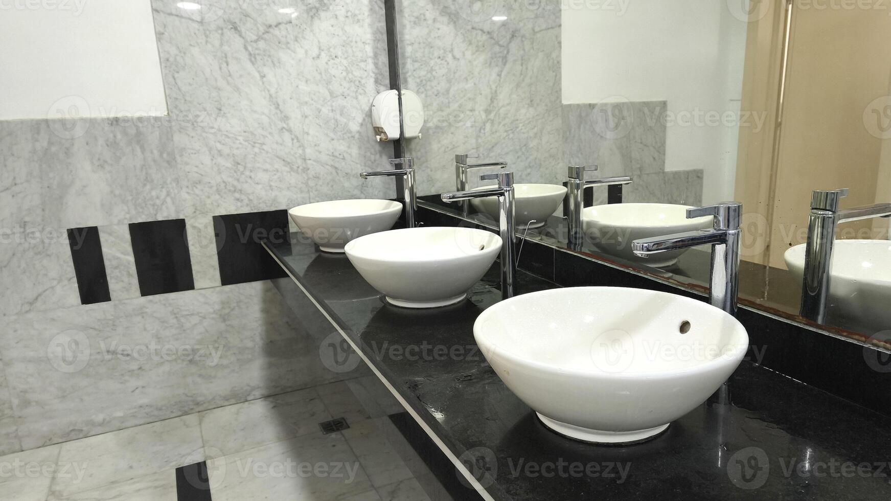 Modern white ceramic sinks with chrome faucets on a black countertop in a clean public restroom, reflected in a wall mirror. Minimalist and functional bathroom interior design. photo