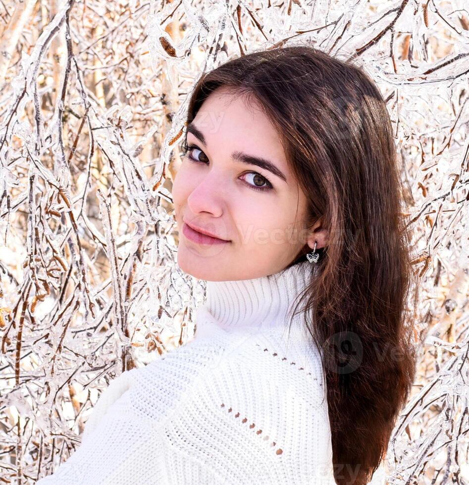 Portrait of a young Russian woman in white sweater looking at camera next to frozen branches of birch trees, covered with ice. Winter forest in Russia photo