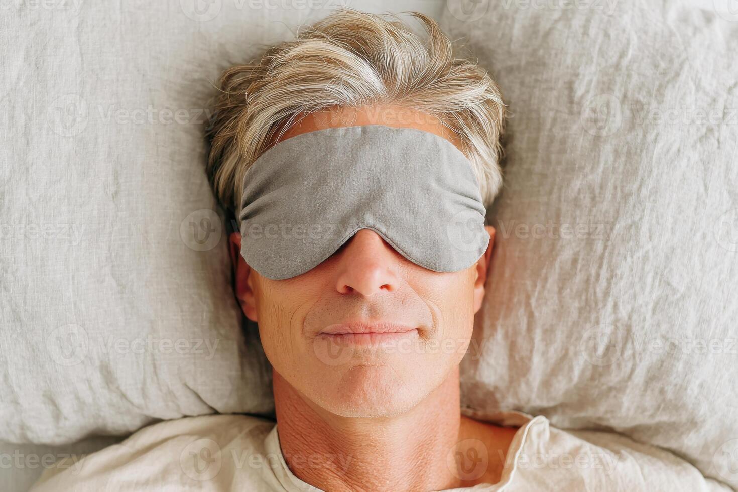 Mature man with gray hair wearing a sleep mask is resting comfortably on a soft pillow, promoting relaxation and self-care in a serene bedroom environment photo