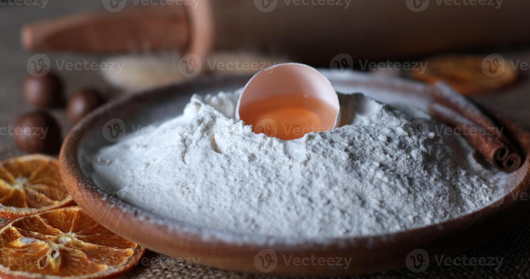 Food still life of organic food ingredients for homemade baking with white flour and egg yolk inside half of broken egg shell. Flour pile in traditional wooden bowl. Dried orange slices and cinnamon photo