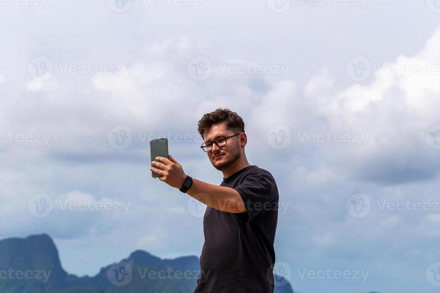 Young man taking selfie outdoors against scenic backdrop of mountains and clouds, showcasing moment of joy and connection with nature and technology. Man taking selfie against sky photo