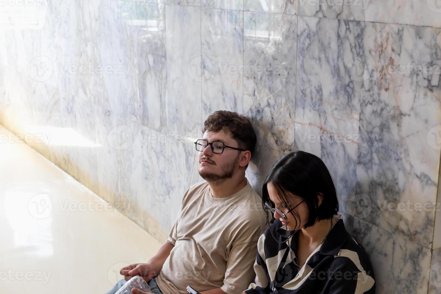 Two tired young adults in glasses are sitting against stylish marble wall. Tired tourists man and woman on tour sitting on floor and resting. Exhaustion and fatigue while traveling photo