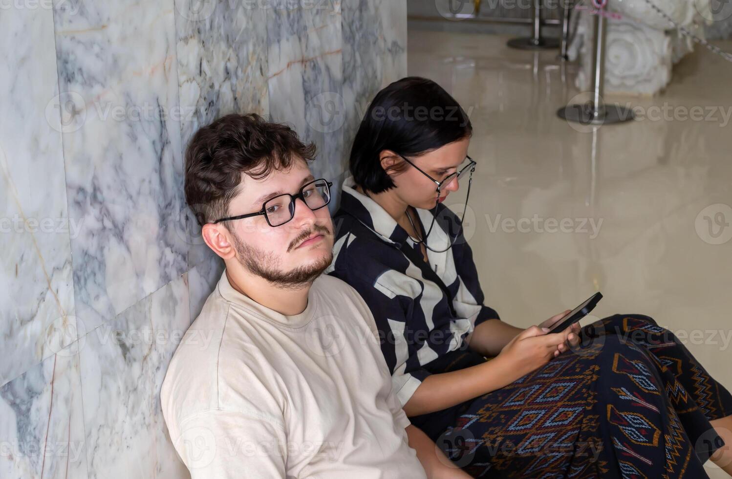 Exhaustion and fatigue while traveling. Two tired young adults are seated on floor against marble wall, one is focused on smartphone while other gazes thoughtfully. Tired tourists sitting on floor photo