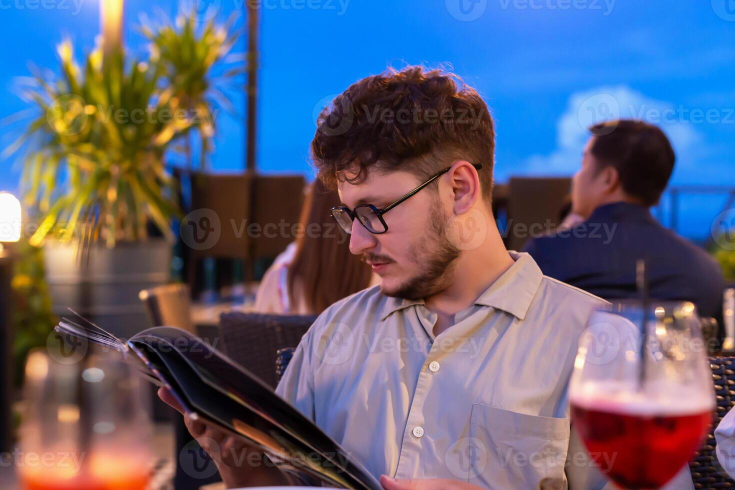 Young man with glasses is focused on reading menu at rooftop restaurant, surrounded by soft lighting and evening ambiance, with drinks and greenery enhancing atmosphere. Restaurant terrace at sunset photo