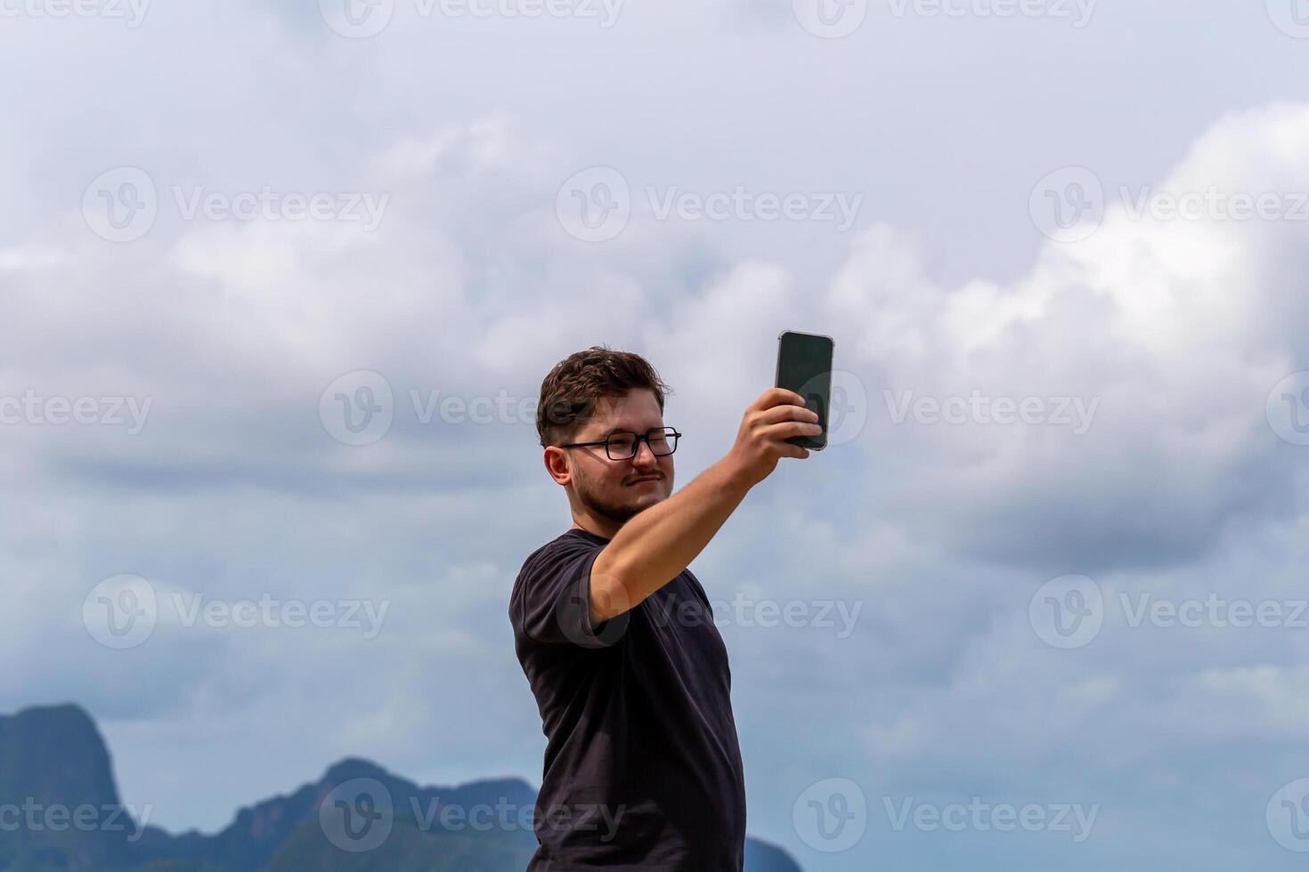 Young man with glasses is taking selfie outdoors against scenic backdrop of mountains and clouds, capturing moment of joy and adventure in nature. Male individual is taking selfie in outdoor setting photo