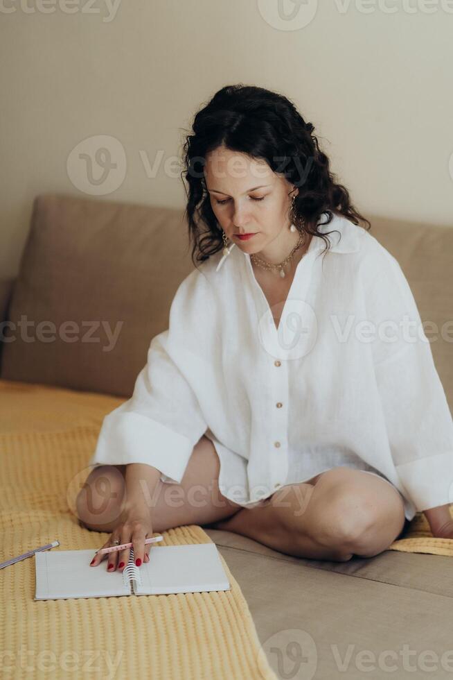 Woman in white linen shirt sits cross legged on beige couch journaling in notebook with pastel pencils in soft light expressing calm and introspective quiet moment of mindful reflection photo