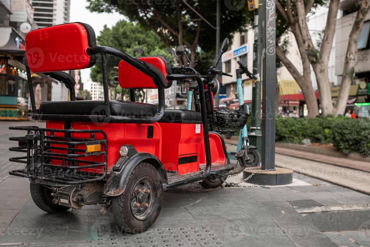 Colorful Three-Wheeled Vehicle Parked on a City Street Surrounded by Greenery and Shops During Daylight Hours photo