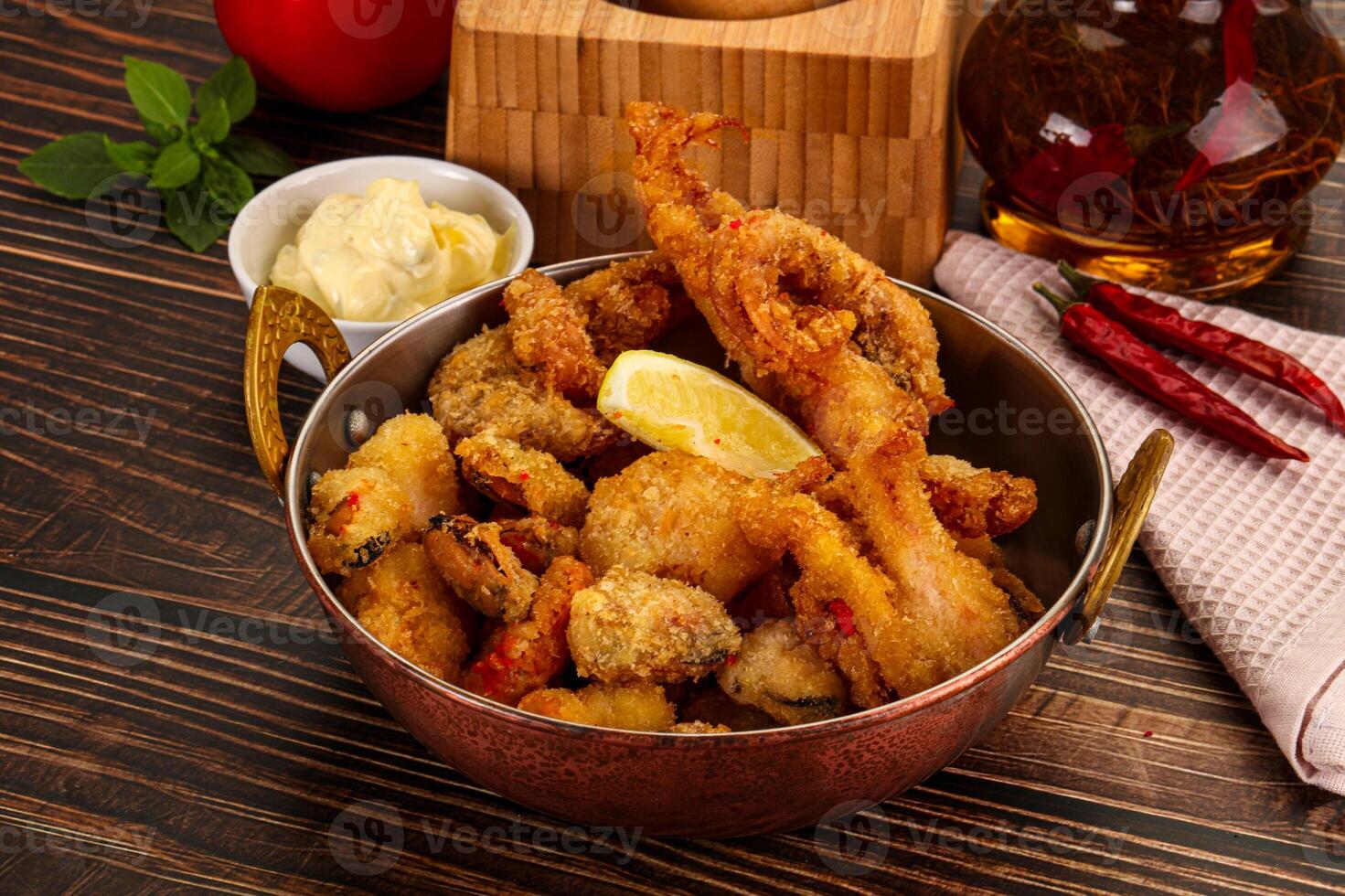 A bowl of fried seafood with lemon wedges and a bottle of sauce photo