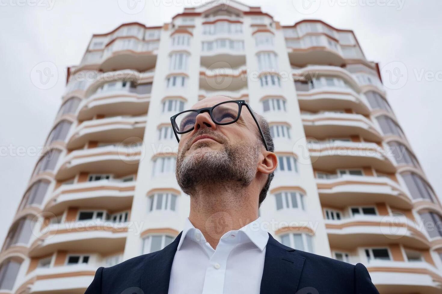 A man in glasses and a suit stands in front of a tall building photo