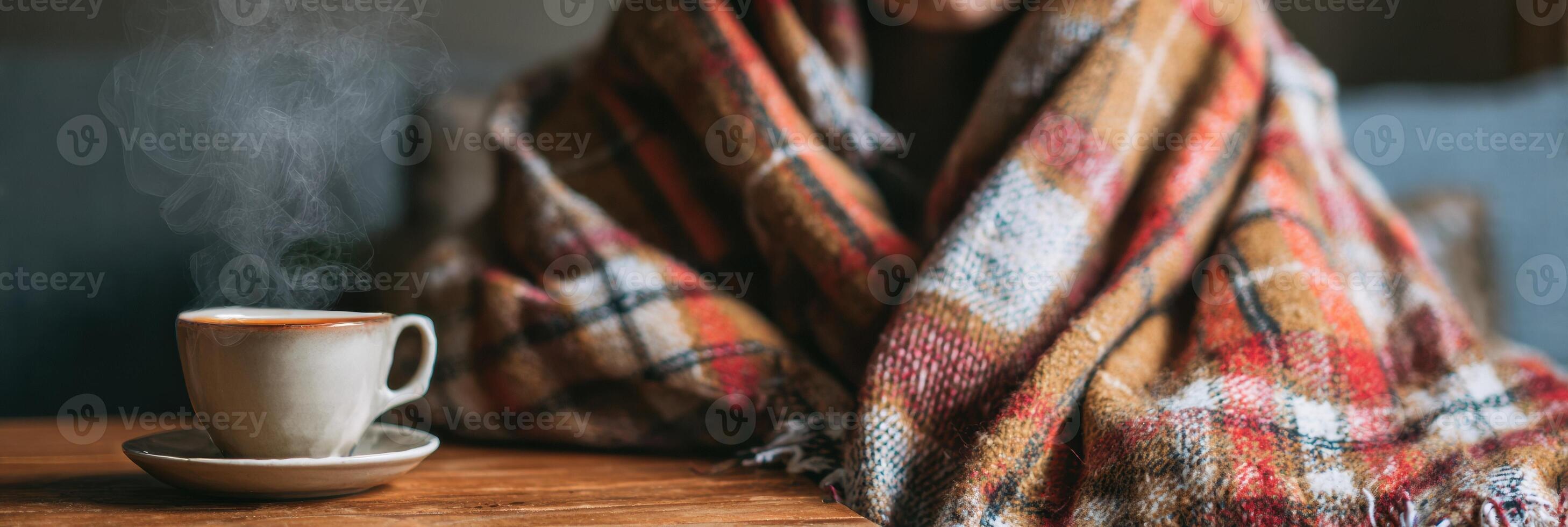 Sick Person Wrapped in Plaid Blanket With Steaming Cup of Tea on Table in Cozy Indoor Setting During Winter Afternoon photo
