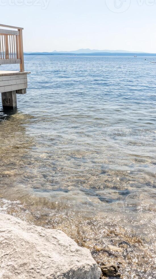 A serene summer day unfolds by the clear, sparkling waters of a tranquil lake, with a rustic wooden dock inviting peaceful contemplation of the distant horizon. photo