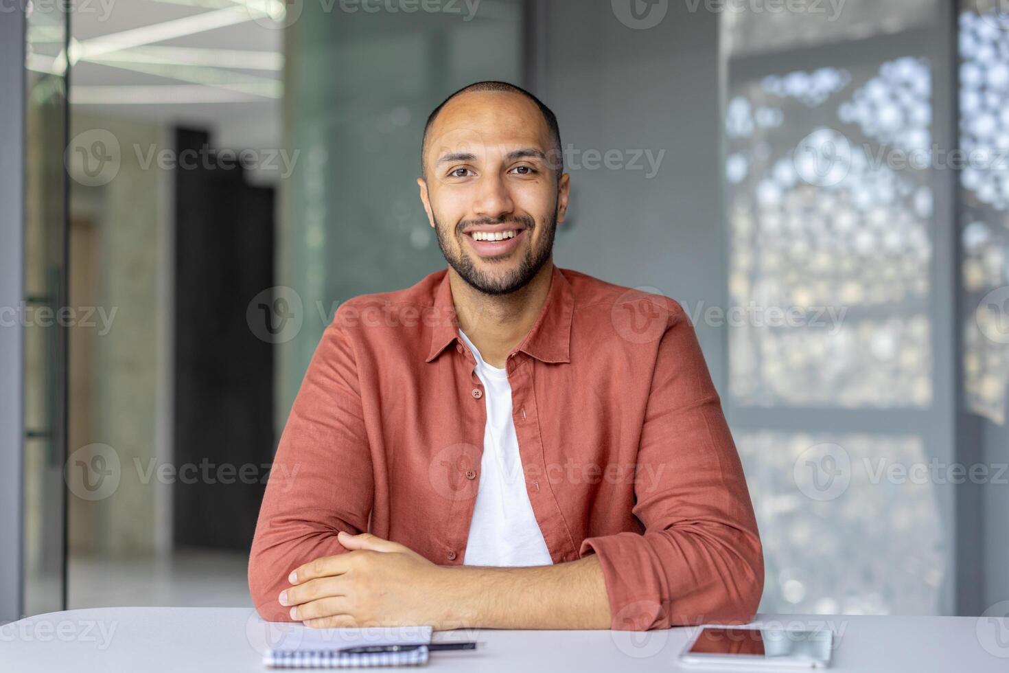 A smiling man sits at a table, appearing confident and relaxed. The setting suggests a modern office or workspace. photo