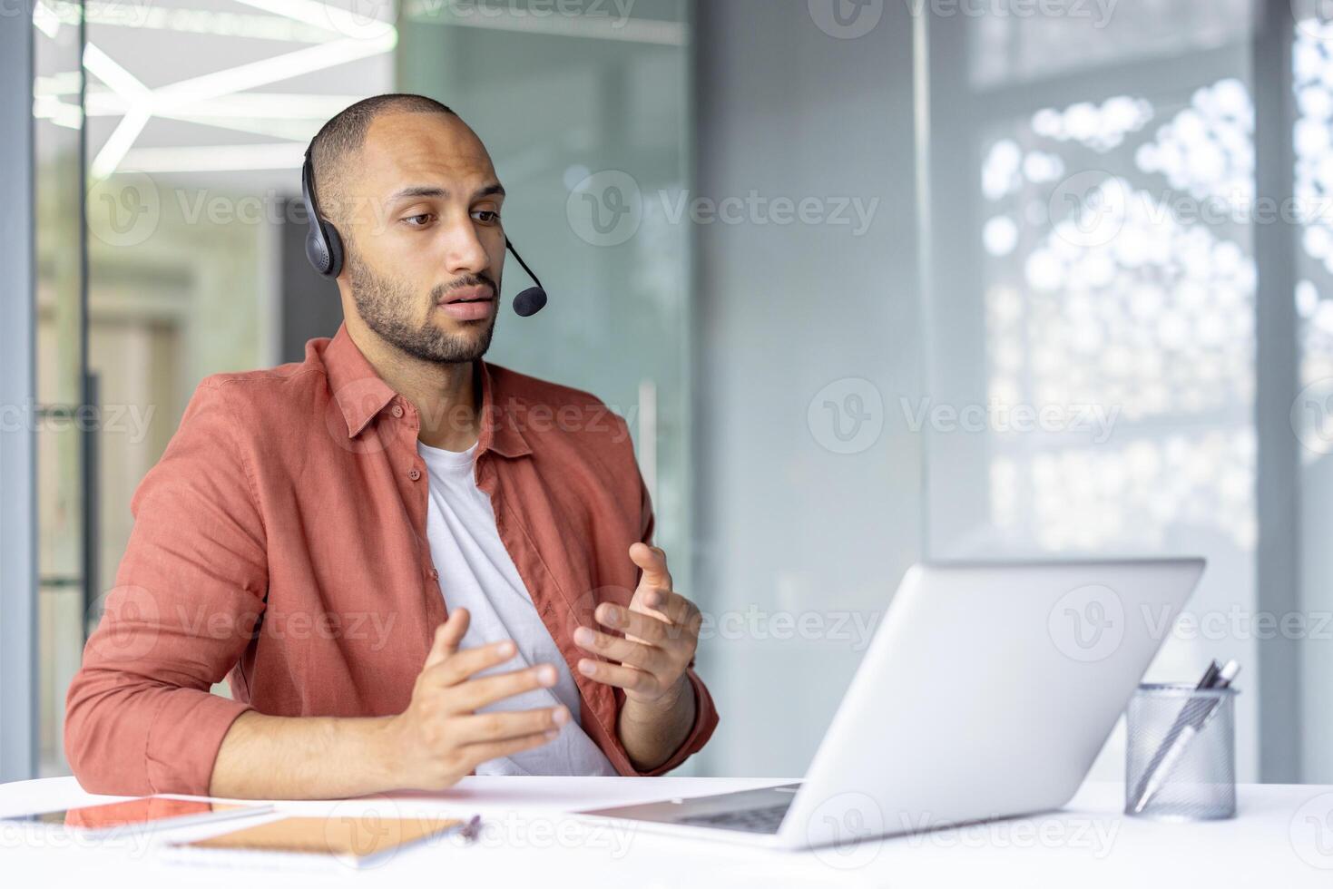 A man in a headset is looking at a laptop and gesturing with his hands during a call in a modern office setting. photo