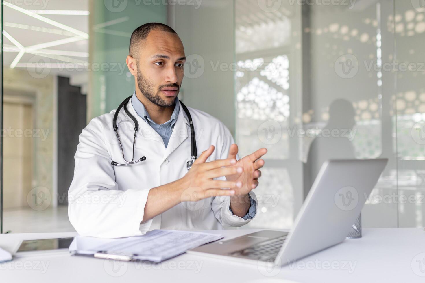 A doctor, wearing a lab coat and stethoscope, gestures while on a call using a laptop in a modern office setting. photo