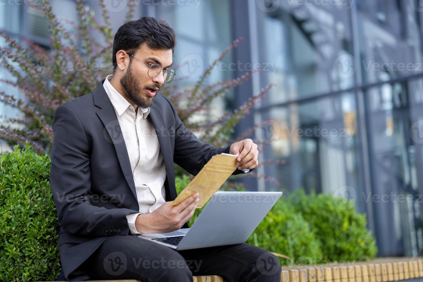 An astonished businessman sits outside with a laptop, examining an envelope with a look of surprise, possibly receiving important documents. photo