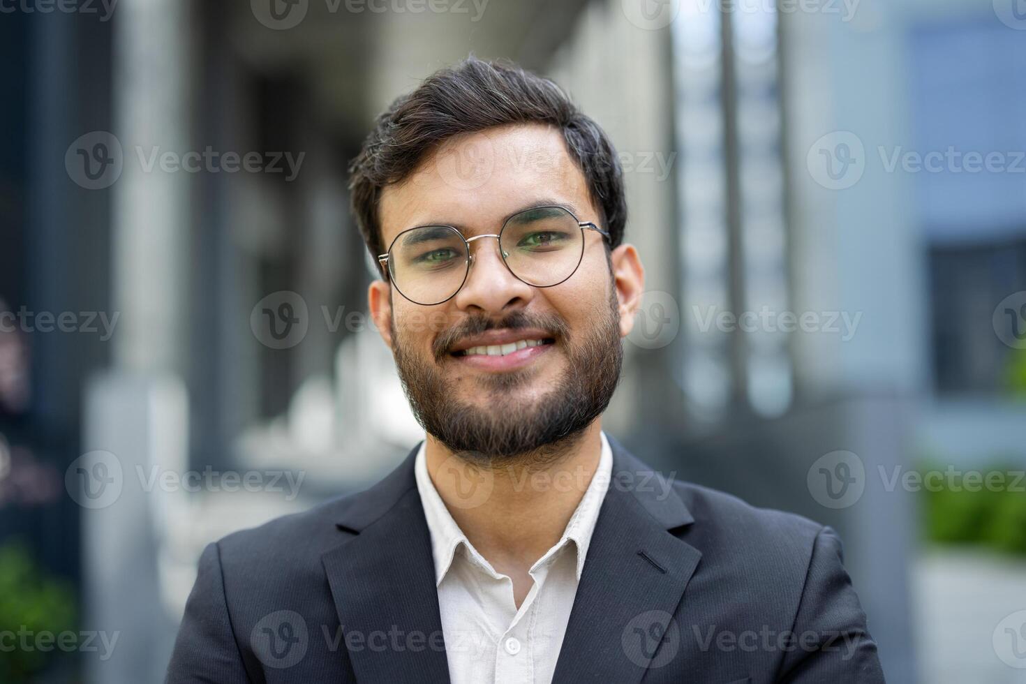 A smiling businessman with a beard and glasses stands confidently outdoors in front of a building, wearing a suit. photo