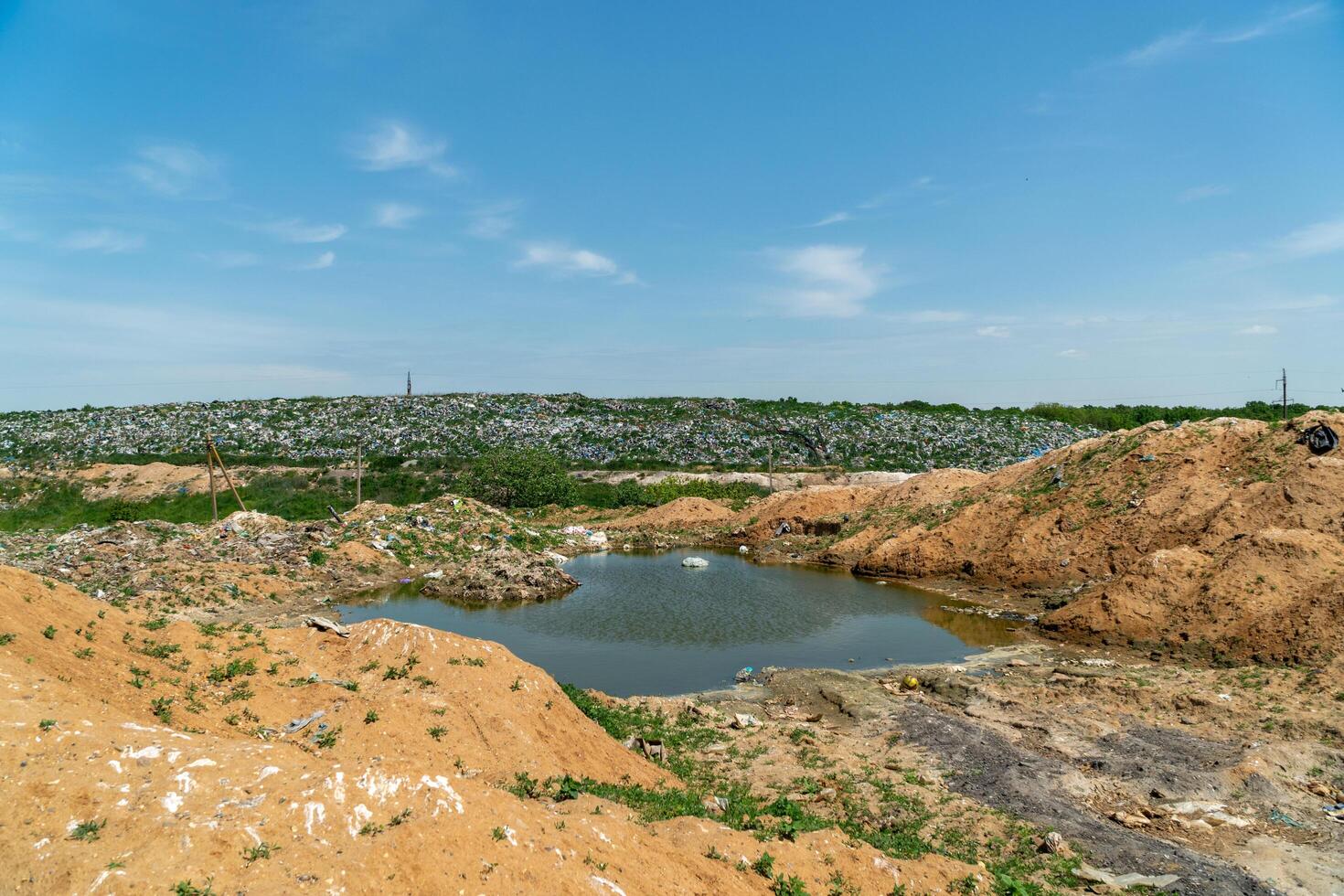 A wasteland of garbage rises in the background with a small pond and patches of greenery under a bright sky photo